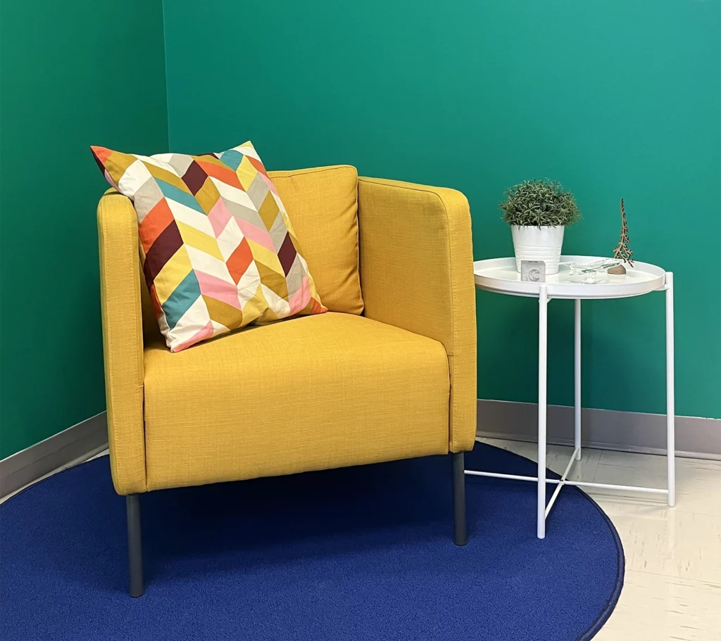 Divan une place jaune de notre agence marketing posé sur un tapis bleu royal rond avec accompagné d'une table d'appoint blanche contenant une plante, le tout sur un fond turquoise.