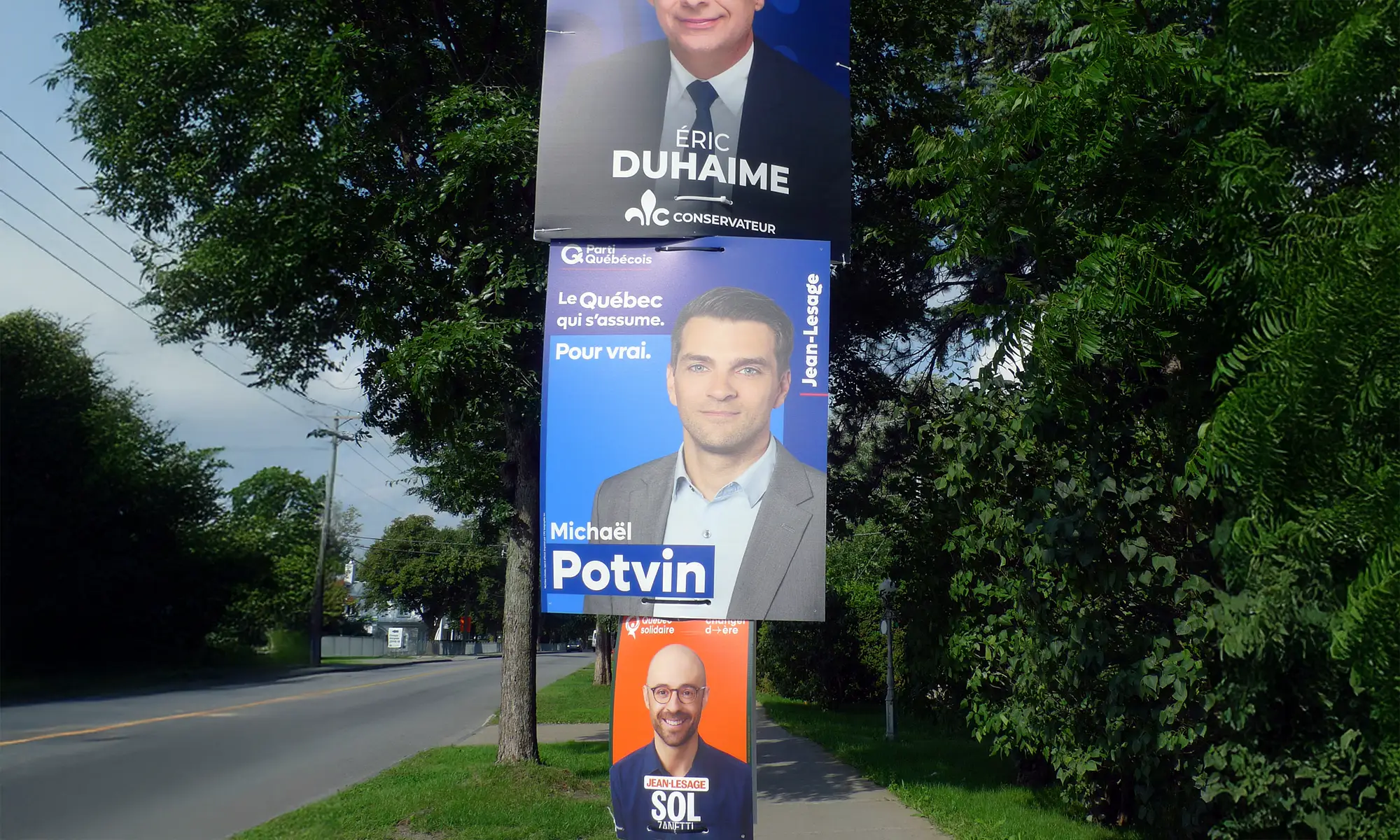 Trois pancartes électorales installées sur un poteau en bord de route, représentant des candidats de différents partis politiques au Québec.
